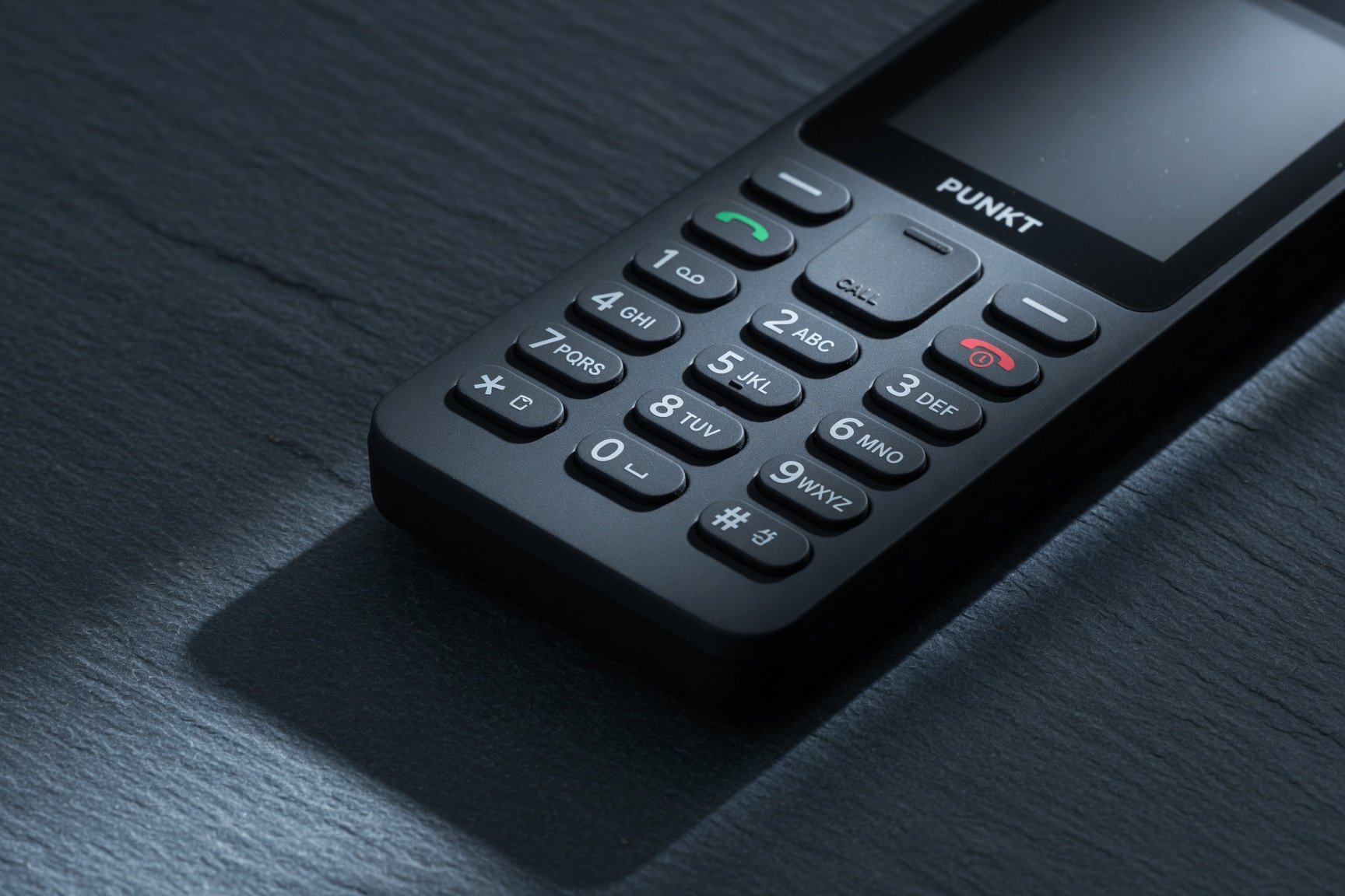 Punkt MP02 matte black minimalist phone on dark slate showing PUNKT branding and T9 keypad